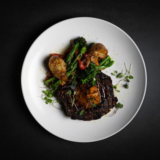 A top view of a grilled steak served with potato, sauce, and greens on a white plate on a black table
