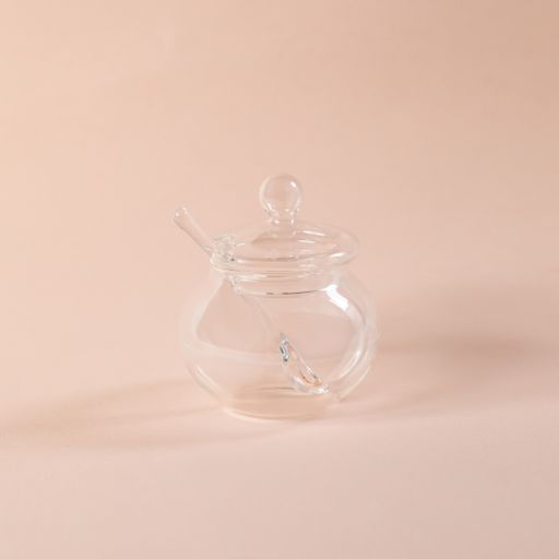 A small decorative glass jar with a lid and a spoon for holding  sugar on a light pink background