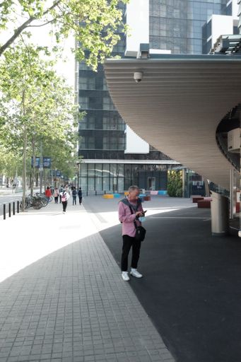 An old man in a pink shirt and sneakers looking at his phone in front of a hotel in Torre Glories area
