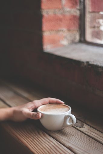 A vertical free-lens shot of a person holding a white coffee cup with cappuccino on a wooden table