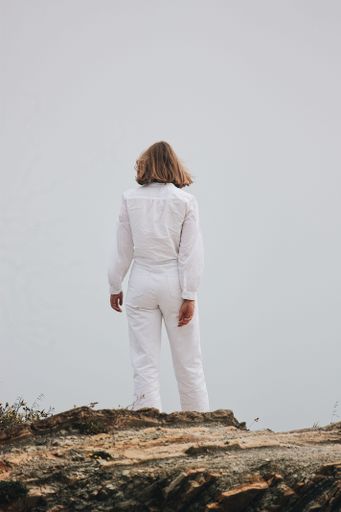 A back view of a young blonde female standing on a cliff in white outfit
