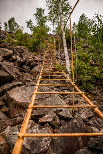 A vertical shot of a yellow metallic bridge above the rocks surrounded by greenery under a cloudy sky