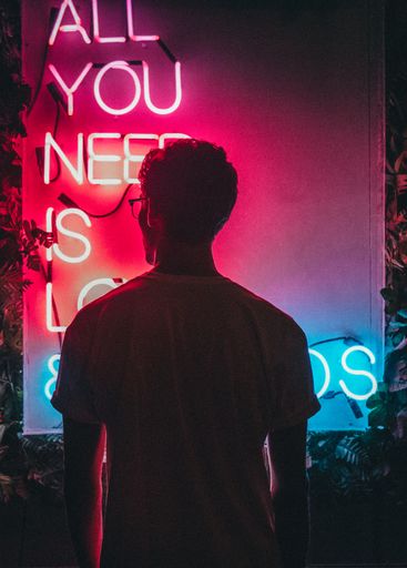 A cool vertical shot of a male standing near "All you need is love" pink and blue neon signage