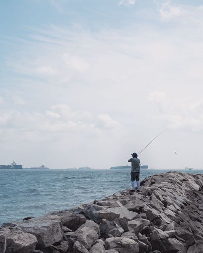 A vertical shot of a person fishing from the rocks of Lazarus island
