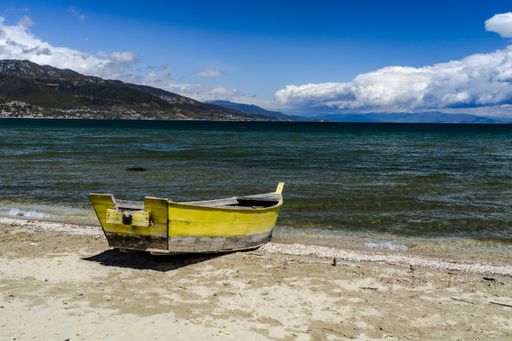 The image shows a yellow rowboat on the shore of the lake which is on the border between Albania and Macedonia.  Mountains and clouds are in distance