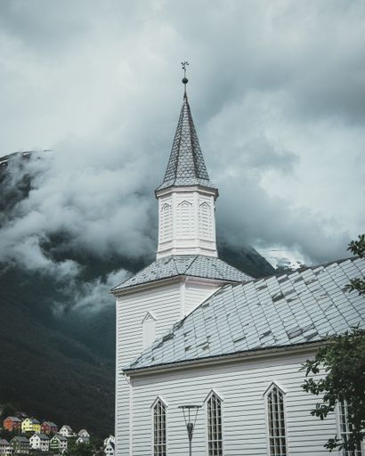 A vertical shot of the white Hyen church in Norway with a mountain hiding behind a cloud in the background