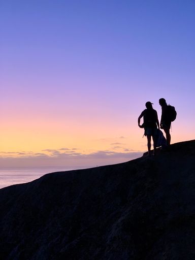 A vertical shot of a silhouette of two persons standing on a hill after long hiking watching the sunset on the horizon
