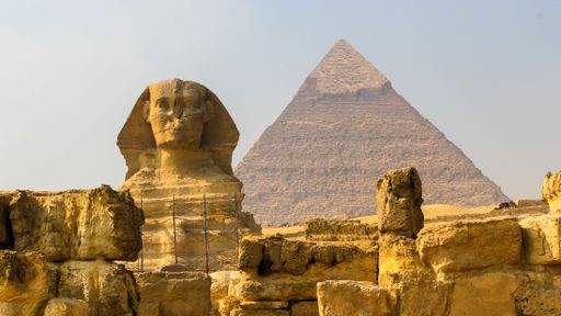 A closeup shot of a Great Sphinx between the ruins with the Cheops Pyramid in the background
