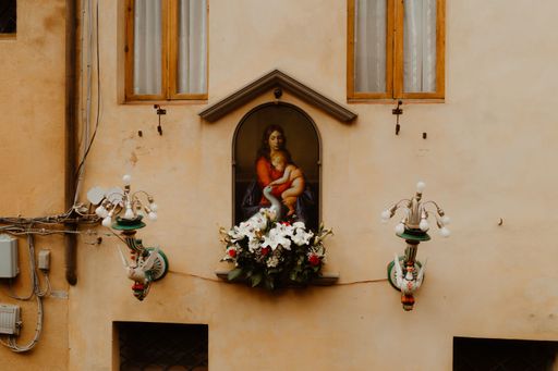 A closeup of a painting of the mother of god decorated with flowers on a building between windows