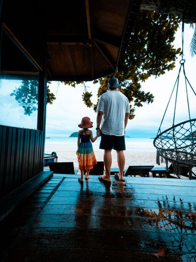 A vertical back view of a father holding the hand of his daughter on a beach resort
