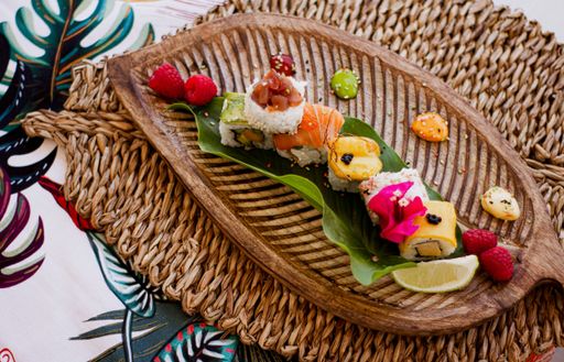 A top view shot of a set of sushi rolls on a wooden leaf-shaped plate with flower and fruit garnish