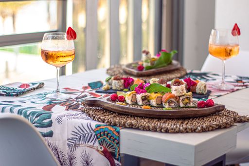 A closeup of sushi sets on plates on a white table with a tropical tablecloth and cups of champagne with a blurred background
