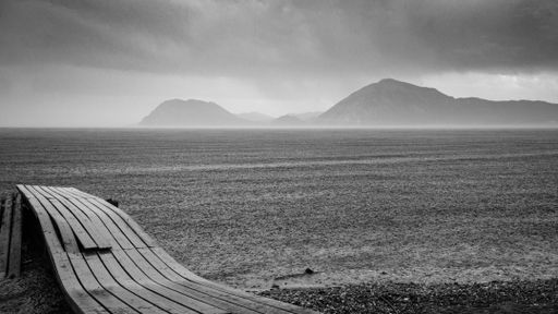 A grayscale of a seaside with a wooden bench on a rainy day with mountains on the other side of the sea