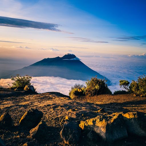 A view of a beautiful volcano at sunrise with clouds around it and a rocky ground in front
