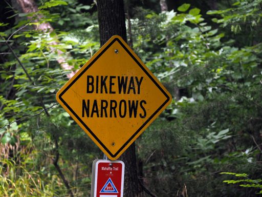 A closeup shot of a yellow sign with the text [BIKEWAY NARROWS] on a wooden pole in a park