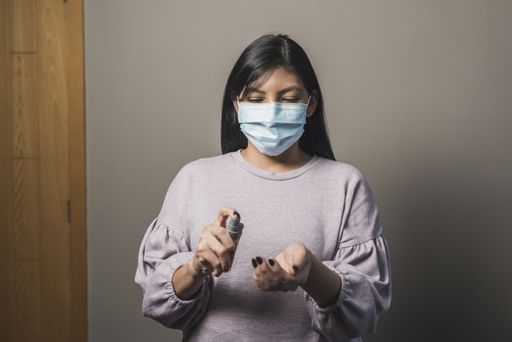 A closeup portrait of a young Hispanic female with long black hair in medical mask applying antibacterial gel on her hands