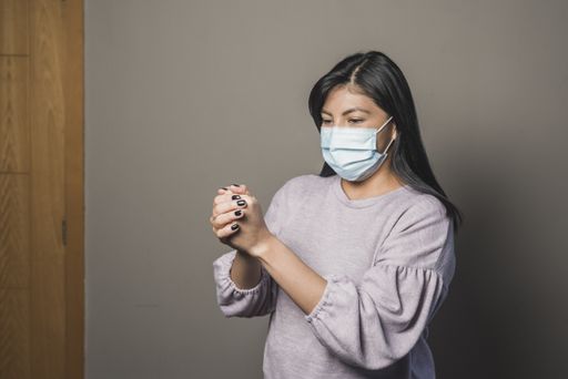 A closeup portrait of a young Hispanic female with long black hair in medical mask applying antibacterial gel on her hands