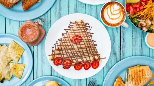 An overhead shot of different dishes including crepes covered with chocolate, salad, a cappuccino, and more on a blue wooden surface