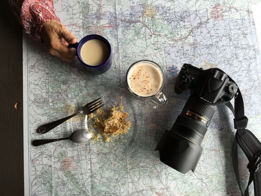 A map, two cups of coffee, a cake, and a camera on a road trip break, top view