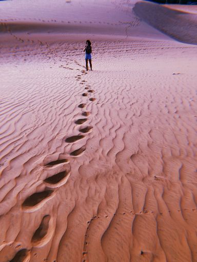 A vertical shot of footprints on sand dunes and a woman standing with a camera