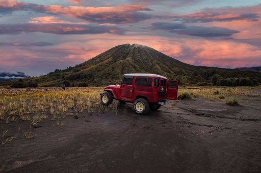 An old red off-road Jeep truck in Indonesia with Mount Bromo under a colorful cloudy sky at sunset