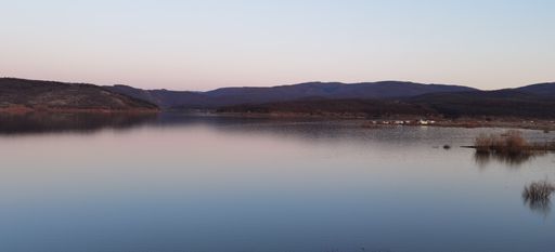 A calm shot of a lake surrounded my flat hills covered in forests under a pink sunset sky