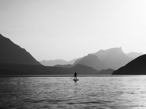 A grayscale shot of a person paddling on a board in the middle of a lake surrounded by lush greenery