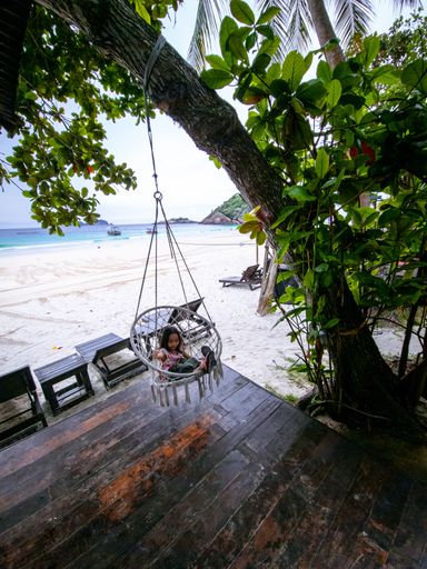 A little girl sitting on the beach swing or hammock with white sand and a calm sea.