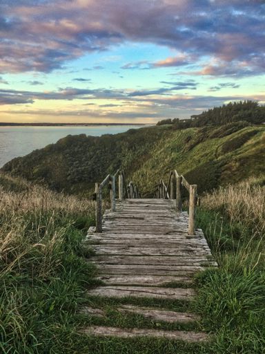 A vertical shot of a wooden dock surrounded by hills covered in greenery and the sea under a cloudy sky in the evening
