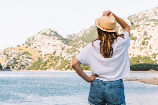 A shallow focus of a young female with a Panama hat surrounded by the sea and hills under the sunlight