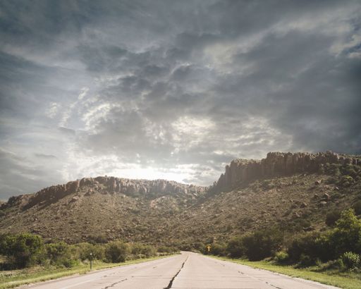 A road surrounded by trees against rocky mountains under the cloudy sky