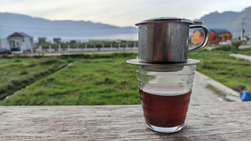 A closeup shot of a glass cup of tea with metal pot on a wooden fence with blurred farm in background