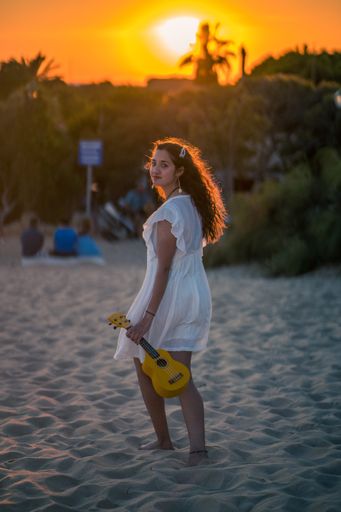 A vertical shot of a young female in a white dress walking on the beach with a ukulele