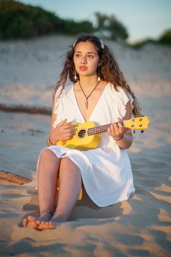 A vertical shot of a young female in a white dress playing ukulele at the beach