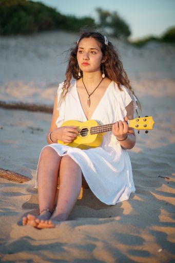 A vertical shot of a pretty European woman in a white light dress playing ukulele while sitting on a sandy beach