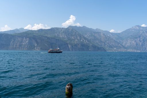 A beautiful shot of a ferry sailing in the Lake Garda in bright sunlight against mountains and blue sky in the background, Italy