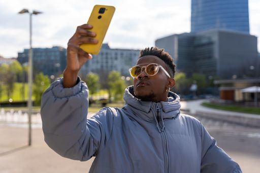 A closeup of a black ethnic man taking a selfie with a phone and a blue jacket in the city