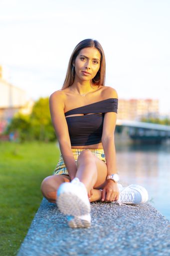 A shallow focus shot of a young Caucasian woman wearing a stylish crop top and yellow checkered skirt, sitting on a rock outdoors during sunset
