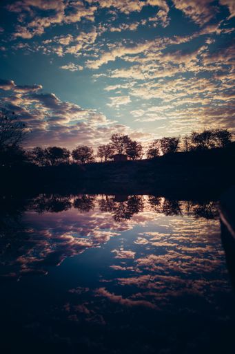 A scenic view of cloudy sky reflecting on pond water with silhouette of trees at sunset