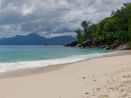 A scenic view of Anse Takamaka beach with takamaka trees on a cliff and mountains in the background