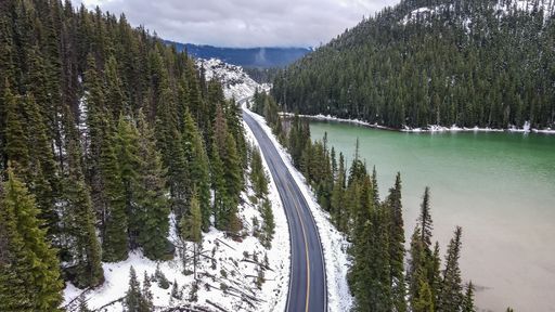 A scenic view of an open road between a lake and mountains with evergreen trees in winter