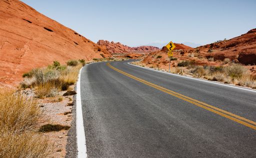 A horizontal shot of an empty highway in the middle of a red rocky desert, with a yellow sign next to the road. Perfect for depicting car travel