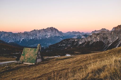 A beautiful shot of a small house near the road and a dry grassy hill with mountains and a clear sky in the background