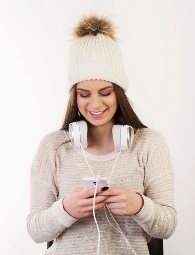 A smiling caucasian female in a sweater and white hat with her headphones and smartphone isolated on a white background