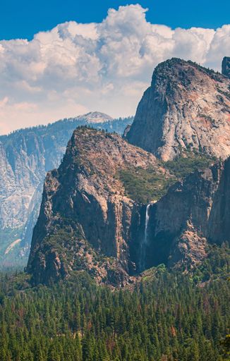 A vertical shot of forested mountains with a waterfall under a blue cloudy sky at daytime