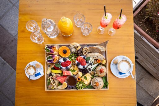 A flat lay of assorted fruits, wraps, and dessert on a platter with drinks and glasses on a wooden table