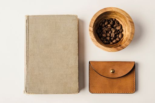 A flat lay of a vintage book,brown leather wallet,and coffee beans in a wooden bowl on white background
