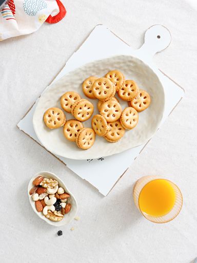 A top view shot of crackers on a white plate, nuts in a smaller bowl and an orange juice