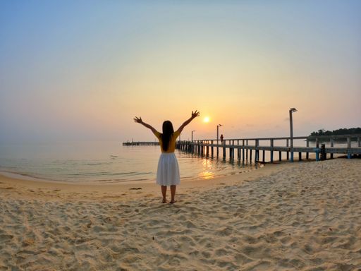 A rear view of a cheerful young woman standing on the sandy beach with hands up enjoying the sunset