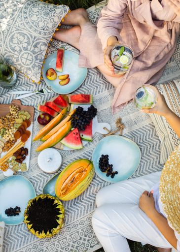 A vertical high angle shot of two friends at a picnic with different fruits and fresh drinks
